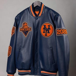 Pode incluir: Um casaco varsity azul-marinho com punhos e gola canelados às riscas laranja e brancas. A frente apresenta um emblema laranja e azul-marinho 'NY', um gráfico de basquetebol 'NWYORK' e o número '2026' na manga com emblemas adicionais.