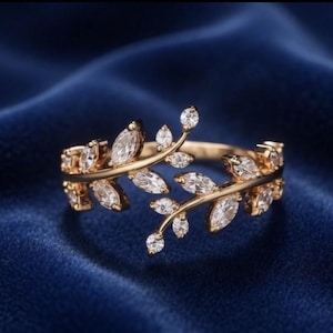 Peut inclure: Une bague dorée avec un motif de feuilles. La bague présente des pierres précieuses claires, de taille marquise, serties le long des feuilles. La bague est présentée sur une surface de velours bleu foncé, mettant en valeur l'éclat et les détails complexes de la bague.