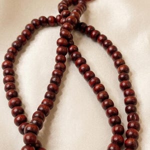 Puede incluir: Un collar de cuentas de madera marrón oscuro con una borla roja. Las cuentas lisas y redondas están ensartadas para formar un collar. La borla está hecha de múltiples hilos rojos.