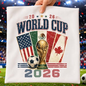 May include: A white fabric item featuring a 2026 World Cup graphic. The design includes the flags of the United States, Mexico, and Canada, a gold trophy, a soccer ball, and the text 2026 WORLD CUP in a distressed, vintage-style font.