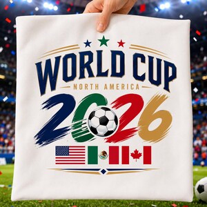 May include: A white fabric square featuring the text WORLD CUP NORTH AMERICA 2026 in dark blue. The number 2026 is stylized with brush strokes in blue, green, red, and gold, with a soccer ball in the center. Below are the flags of the USA, Mexico, and Canada.