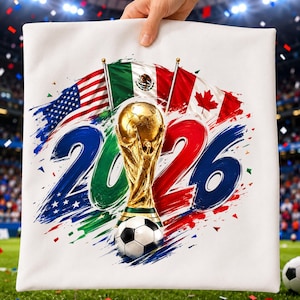 May include: A white square pillow featuring a graphic of the year 2026 in blue and red brushstrokes, a gold soccer trophy, a black and white soccer ball, and the flags of the United States, Mexico, and Canada arranged in a fan shape above the text.