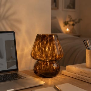 Puede incluir: Una lámpara con forma de hongo de cristal marrón se encuentra sobre un escritorio de madera junto a un portátil y libros. La lámpara tiene un patrón texturizado y emite un brillo cálido. Es una lámpara decorativa.