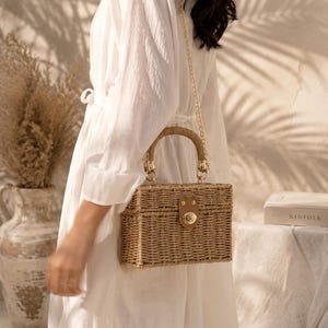 Puede incluir: Bolso de mano rectangular de ratán tejido con una correa de cadena dorada y un cierre de cuero marrón. El bolso tiene un asa redondeada. La persona lleva un vestido blanco. El fondo incluye un jarrón y un libro titulado "KINFOLK".