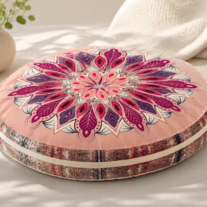 May include: A round floor cushion featuring a light pink top surface with a large, symmetrical mandala pattern in shades of magenta, purple, and white. The side of the cushion has a textured, multi-colored patterned border with a cream-colored trim.
