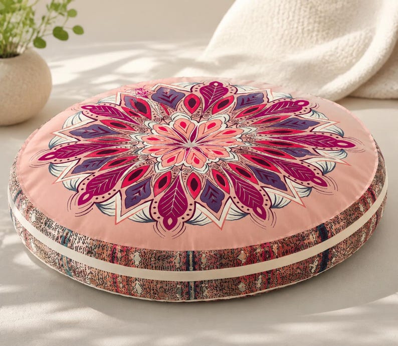 May include: A round floor cushion featuring a light pink top surface with a large, symmetrical mandala pattern in shades of magenta, purple, and white. The side of the cushion has a textured, multi-colored patterned border with a cream-colored trim.