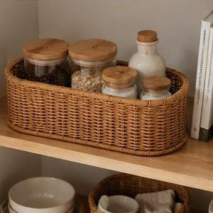 May include: A woven wicker basket filled with glass jars and a white ceramic bottle with wooden lids. The basket sits on a wooden shelf, alongside a stack of books. The jars contain grains and other dry goods.