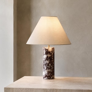 Peut inclure: Une lampe de table avec une base cylindrique en pierre marbrée blanche et violet foncé. La lampe est surmontée d'un abat-jour conique en tissu blanc cassé diffusant une lueur douce et chaude, soutenu par une fine tige centrale dorée.