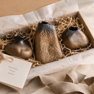 Peut inclure: Un coffret cadeau contenant trois vases en céramique faits à la main, de couleurs marron et noir. Les vases ont des formes et des textures variées, et sont nichés dans du papier et du rembourrage décoratif. Une carte indique "Handcrafted, Natural, Timeless".