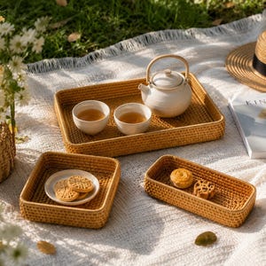 Puede incluir: Escena de picnic con bandejas de ratán tejido. Una bandeja rectangular contiene una tetera y dos tazas de té. Una bandeja cuadrada contiene galletas y una bandeja larga contiene pasteles.