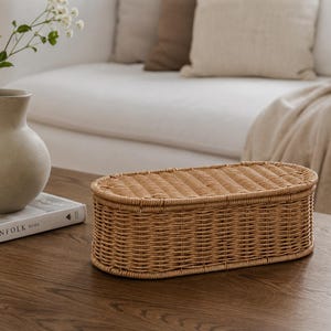 May include: A woven rattan storage basket with a lid, oval in shape, sits on a dark wood surface. The basket is light brown and has a textured surface. A vase with flowers and a book are in the background.