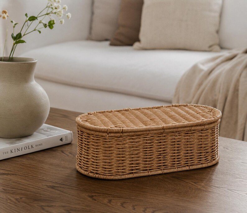 May include: A woven rattan storage basket with a lid, oval in shape, sits on a dark wood surface. The basket is light brown and has a textured surface. A vase with flowers and a book are in the background.