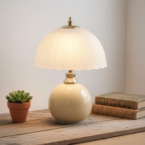 Peut inclure: Une lampe de table crème avec un abat-jour festonné et une base sphérique. La lampe a des accents en laiton et repose sur une surface en bois. Une petite plante succulente en pot et deux livres sont également sur la table.