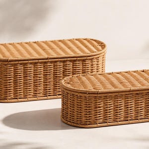 May include: Two woven wicker storage baskets in a light brown color. The baskets are oval-shaped with matching lids. The larger basket is in the background, and the smaller one is in the foreground. The baskets are on a white surface.