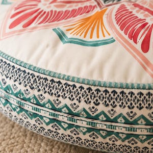 May include: A round floor cushion featuring a cream-colored top with a large, symmetrical floral-inspired pattern in shades of red, orange, and teal. The side panel displays a detailed, repeating geometric print in dark blue and teal.