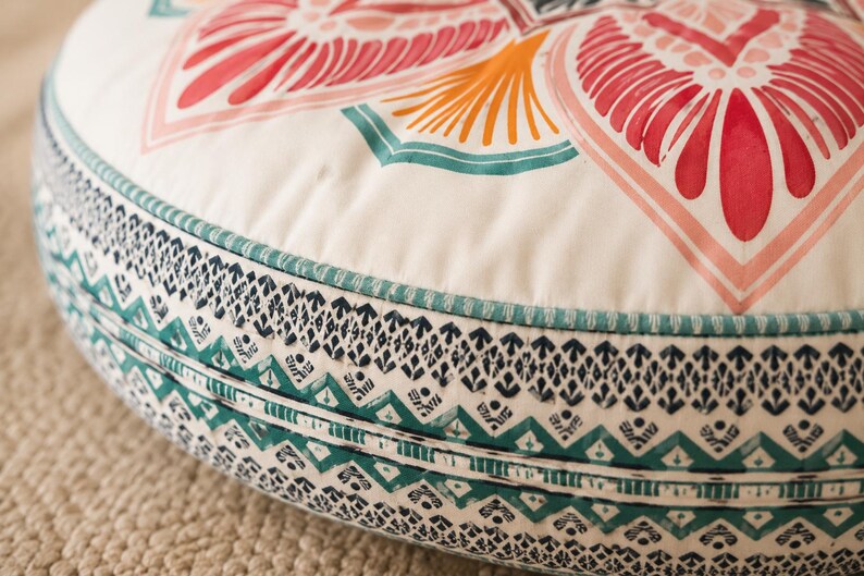 May include: A round floor cushion featuring a cream-colored top with a large, symmetrical floral-inspired pattern in shades of red, orange, and teal. The side panel displays a detailed, repeating geometric print in dark blue and teal.