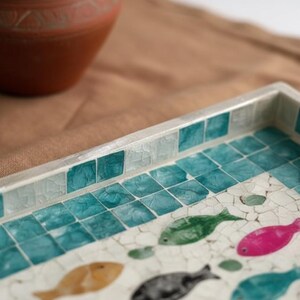 May include: Decorative rectangular tray with a mosaic tile design featuring teal squares and colorful fish. The tray has a silver border and is set on a brown surface. A terracotta-colored vase is in the background.
