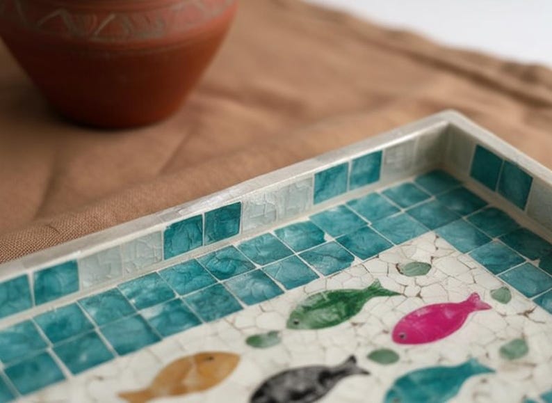 May include: Decorative rectangular tray with a mosaic tile design featuring teal squares and colorful fish. The tray has a silver border and is set on a brown surface. A terracotta-colored vase is in the background.