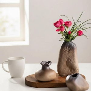 Peut inclure: Nature morte avec une tasse blanche, un plateau en bois et trois vases en céramique. Un vase contient des roses roses. Les vases sont dans des tons de marron et de gris.
