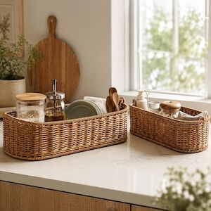 May include: Two woven rattan baskets on a countertop. One basket holds plates, utensils, and a glass jar. The other basket contains a jar, a bottle, and a cloth. A wooden cutting board and a plant are in the background.