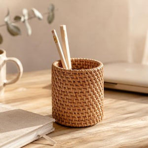 Puede incluir: Un portalápices cilíndrico de ratán tejido, con dos lápices dentro. El soporte marrón claro está sobre un escritorio de madera, junto a una taza beige y un cuaderno cerrado. La escena está bañada por la cálida luz del sol.