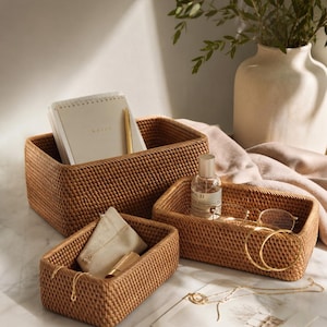 Peut inclure: Trois paniers de rangement en rotin tressé de différentes tailles. Le plus grand contient un carnet et un stylo. Le moyen contient un flacon de parfum et des lunettes. Le plus petit contient une pochette et des bijoux. Tous les paniers sont de couleur marron chaud.