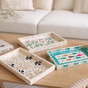 Op de afbeelding: Drie decoratieve rechthoekige trays met mozaïekontwerpen. Eén tray heeft een bloemenpatroon, een andere heeft een vismotief in turquoise en de derde toont groene planten. De trays staan op een houten tafel.
