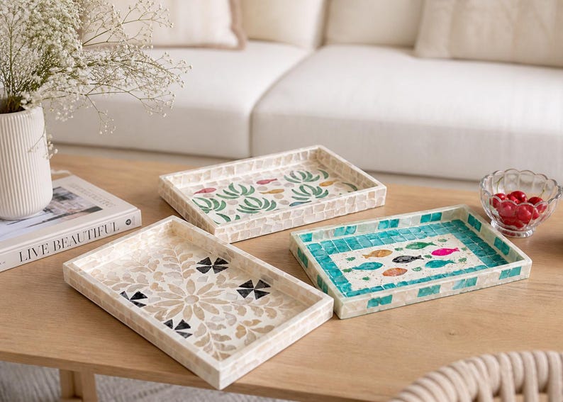 May include: Three decorative rectangular trays with mosaic designs. One tray features a floral pattern, another has a fish motif in turquoise, and the third displays a green plant design. The trays are on a wooden table.