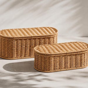 May include: Two woven wicker storage baskets in a natural tan color. The baskets are oval-shaped with matching lids. The larger basket is approximately 24 inches long, and the smaller one is about 16 inches long. The baskets are suitable for home organization.