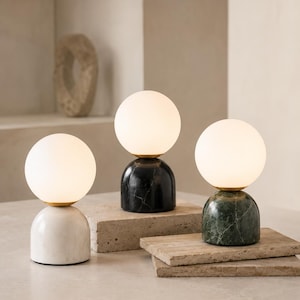Peut inclure: Trois lampes de table composées d'abat-jours sphériques en verre blanc sur des bases en forme de dôme. Une base est en marbre blanc, une en marbre noir et une en marbre vert. Chaque lampe possède un connecteur en métal doré entre l'abat-jour et la base.