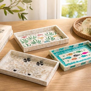 May include: Three decorative rectangular trays with mosaic designs. One tray features a floral pattern, another has a teal and white fish design, and the third has a green and red plant design. The trays are on a wooden surface.