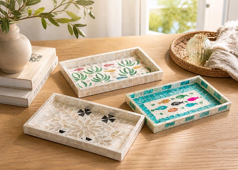 May include: Three decorative rectangular trays with mosaic designs. One tray features a floral pattern, another has a teal and white fish design, and the third has a green and red plant design. The trays are on a wooden surface.