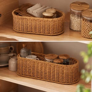 May include: Two wicker baskets, one on each shelf, filled with kitchen items. The top basket holds wooden utensils and folded linen napkins. The bottom basket contains mugs and small jars. Glass jars with wooden lids are also visible.