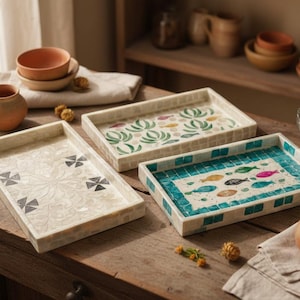 May include: Three decorative rectangular trays with mosaic designs. One tray features a floral pattern, another has a fish motif, and the third has a geometric design. The trays are on a wooden surface.