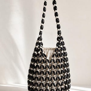 Peut inclure: Un sac seau composé d'un treillis de perles noires allongées et de petites perles blanches sphériques. Le sac présente une longue anse réalisée avec le même motif de perles et comprend une pochette intérieure en tissu blanc cassé avec cordon.