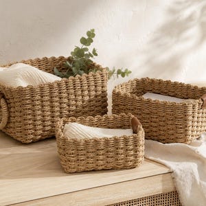 Peut inclure: Trois paniers rectangulaires tressés de différentes tailles. Les paniers sont de couleur marron clair avec un bord festonné. Le plus grand a des poignées rondes, les autres ont des accents en cuir. Des serviettes blanches sont à l'intérieur.