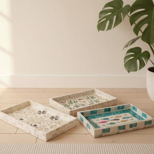 May include: Three decorative rectangular trays with mosaic tile designs. One tray features a floral pattern, another has a botanical design, and the third showcases colorful fish. The trays are on a light wood surface.