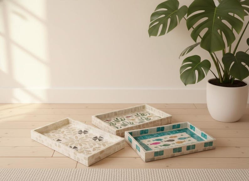 May include: Three decorative rectangular trays with mosaic tile designs. One tray features a floral pattern, another has a botanical design, and the third showcases colorful fish. The trays are on a light wood surface.