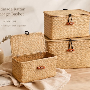 May include: Handmade rattan storage baskets with lids. The rectangular baskets are woven in a natural beige colour. One basket is open, revealing the interior. The lids are secured with a black cord and a wooden toggle. The text on the image says "Handmade Rattan Storage Basket With Lid".
