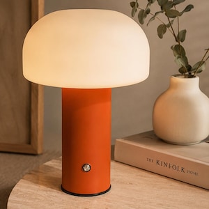 May include: An orange table lamp with a white, mushroom-shaped shade. The lamp has a cylindrical base and a small, round button. The lamp is on a light-coloured, round table. A vase and a book are in the background.