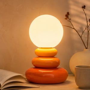 Könnte beinhalten: Moderne orange-gelbe Tischlampe mit einem gestapelten, abgerundeten Sockel und einem kugelförmigen Licht. Die Lampe ist ein dekoratives Akzentstück mit warmem Glanz. Die Lampe ist etwa 30 cm hoch.