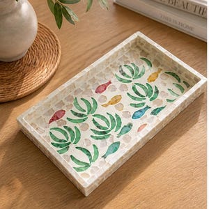 May include: Rectangular decorative tray with a mosaic tile design. The tray features a white border and a mosaic pattern of green leaves and colorful fish. The tray is on a wooden surface, with a vase and books in the background.