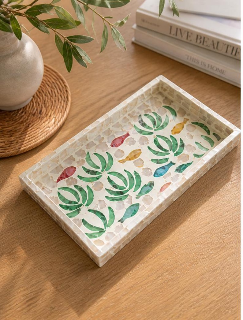 May include: Rectangular decorative tray with a mosaic tile design. The tray features a white border and a mosaic pattern of green leaves and colorful fish. The tray is on a wooden surface, with a vase and books in the background.