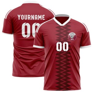 Puede incluir: Camiseta de f&uacute;tbol granate con detalles blancos, con el n&uacute;mero "00" en blanco en la parte delantera y trasera. La camiseta tiene cuello en V y un patr&oacute;n en zigzag en el centro. La espalda muestra "YOURNAME". Un logo del equipo est&aacute; en el pecho.