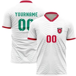 Può includere: Maglia da calcio bianca con rifiniture rosse e scollo a V. La maglia presenta il numero "00" in rosso sul davanti e sul retro, oltre al testo "YOURNAME" e al numero "00" in verde sul retro. Un emblema della Giordania è sul davanti.