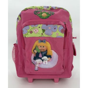 May include: A pink rolling backpack featuring a Cabbage Patch Kids logo at the top and a light green panel with a purple and pink floral pattern. The front pocket displays a graphic of a blonde doll holding a green phone with a small white dog.