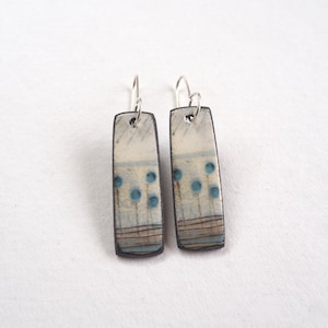 May include: A pair of dangle earrings with a rectangular ceramic charm featuring a white background with blue and brown abstract designs.