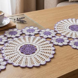 Puede incluir: Un camino de mesa de ganchillo hecho a mano con una serie de motivos circulares interconectados. El diseño consta de dos tapetes grandes de color blanco y morado rodeados de pequeños detalles en forma de flor con hilos de colores a juego.