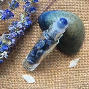 Puede incluir: Un frasco de aceite esencial con rodillo de vidrio transparente lleno de piedras azules. El frasco tiene una tapa de bola rodante azul. Una concha azul y flores azules están en el fondo. Los artículos están sobre una superficie tejida marrón.