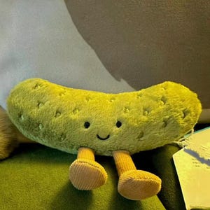 Op de afbeelding: Een pluche augurk-speeltje in een levendige limoengroene kleur met een zachte, pluizige textuur. De augurk heeft een eenvoudig geborduurd gezicht met twee zwarte ogen en een kleine gebogen glimlach. Het heeft twee korte, lichtbruine ribfluwelen benen.
